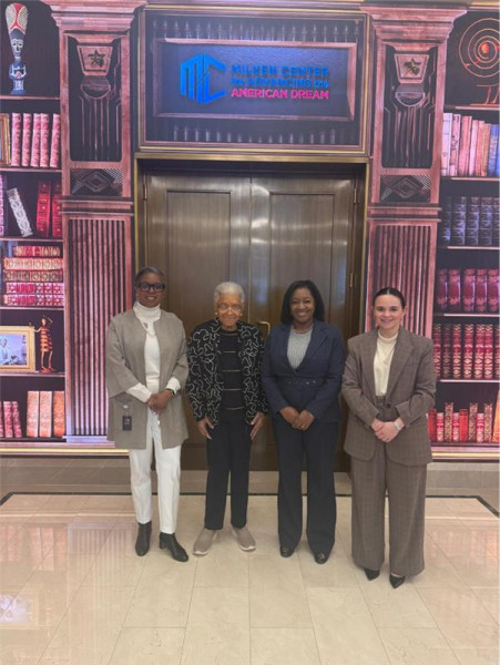 Four people standing together in formal attire in front of a television and pink bookshelves